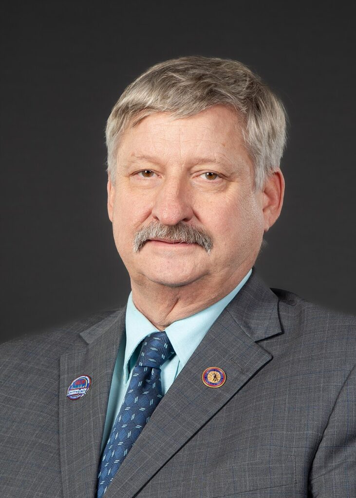A portrait of a man with gray hair and a mustache wearing a gray suit, a light blue shirt, and a blue patterned tie. He has two pins on his suit jacket.