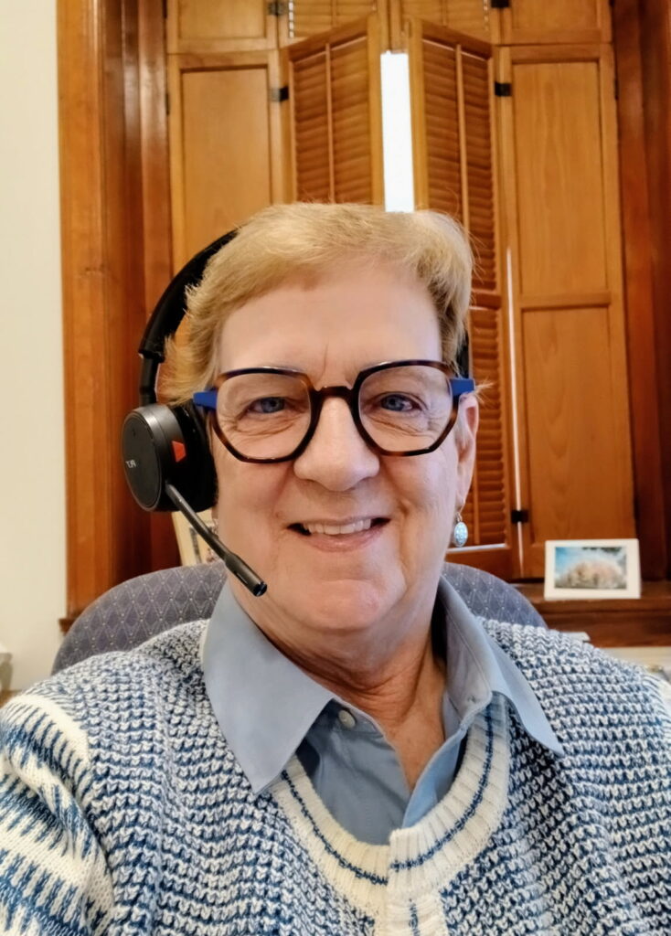 A woman with blonde hair and glasses smiles while wearing a headset and a blue and white patterned sweater over a light blue collared shirt.