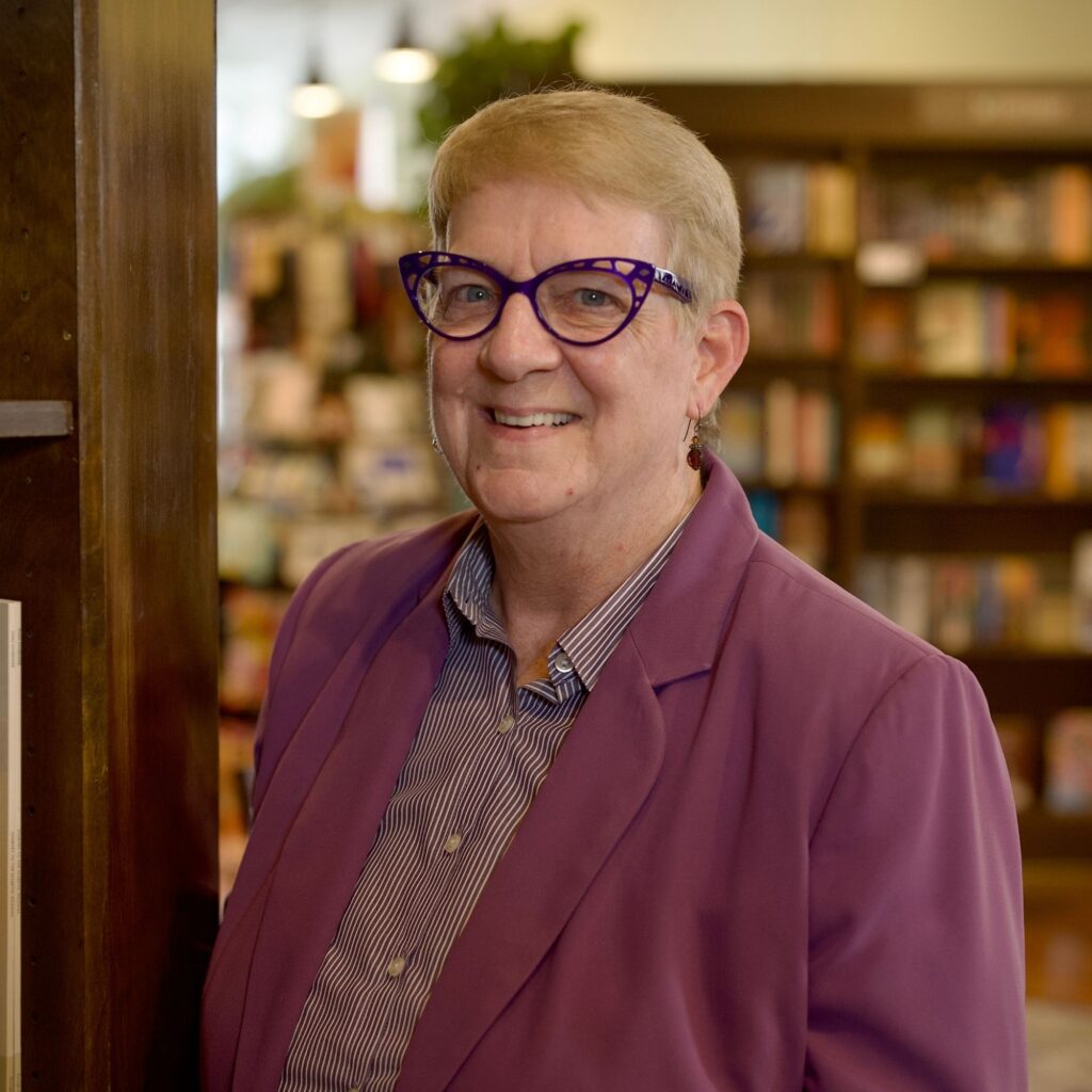 A woman with short blonde hair and purple cat-eye glasses smiles at the camera. She is wearing a purple blazer over a striped shirt and stands in front of bookshelves.