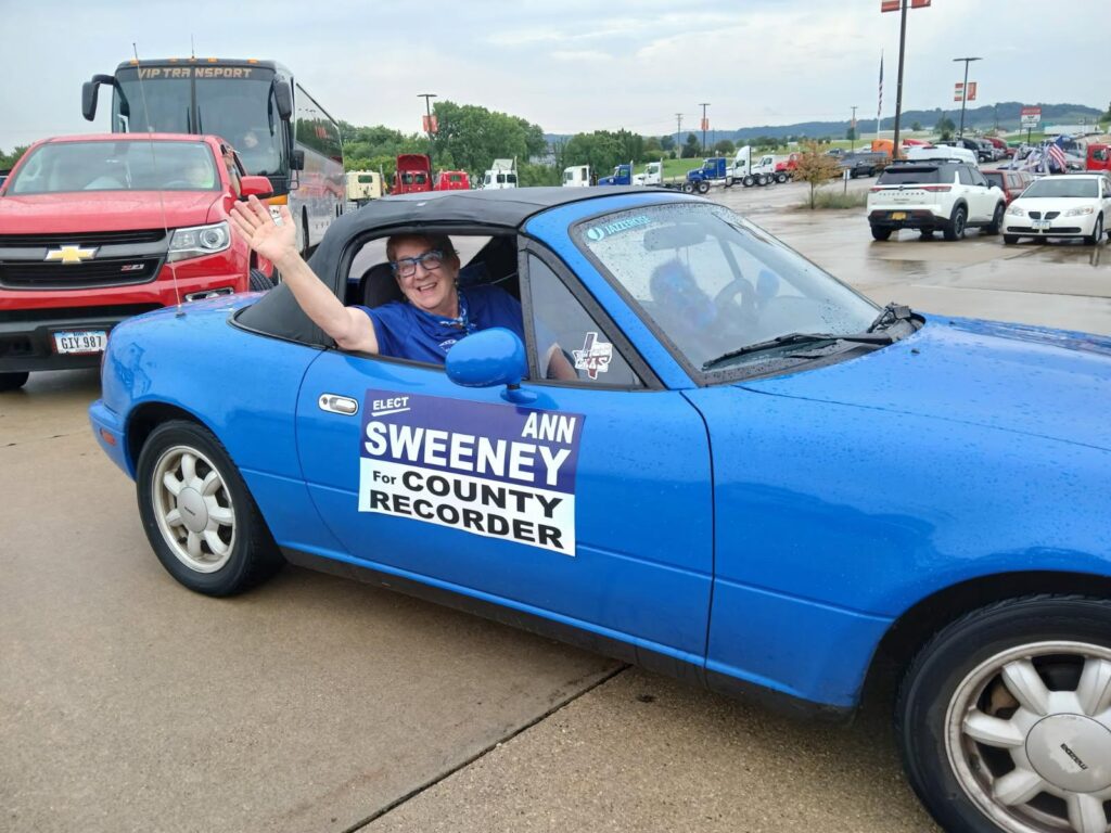 A woman in a blue convertible waves from the driver's seat. A large sticker on the car reads "ELECT ANN SWEENEY For COUNTY RECORDER".