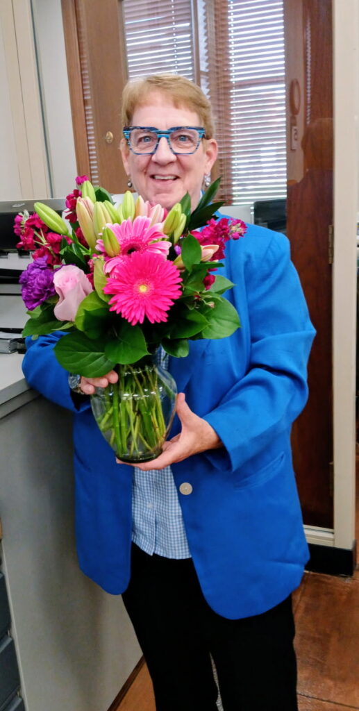 A woman with short blonde hair and blue-rimmed glasses smiles as she holds a vase of colorful flowers, including pink gerbera daisies and lilies, while wearing a bright blue blazer.