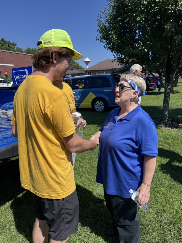 A man wearing a yellow t-shirt and a neon yellow cap with "Manwich" written on it shakes hands with a woman wearing a blue polo shirt and sunglasses. They are standing on a grassy lawn on a sunny day, with a blue van in the background that has "dubcolib.org" and "County District" written on it.
