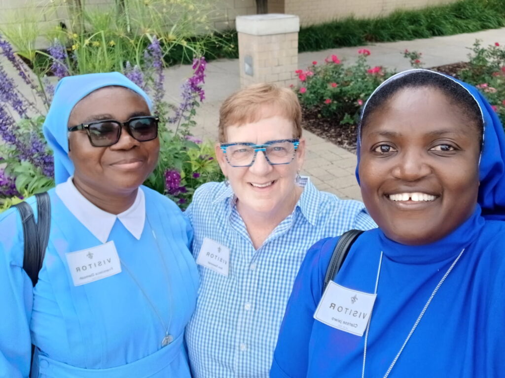 Three women in blue habits and a woman in a blue and white checkered shirt smile at the camera in front of a garden.