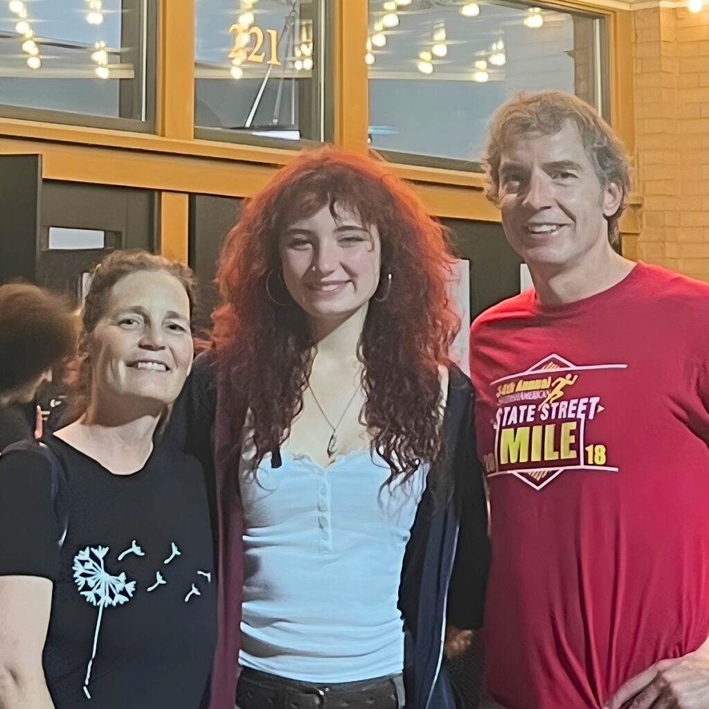 A smiling family of three poses for a photo in front of a building with "221" visible on the facade. The woman on the left wears a black t-shirt with a dandelion graphic, the young woman in the center has long, curly red hair and wears a white tank top, and the man on the right wears a red t-shirt that reads "34th Annual America's State Street Mile 18".