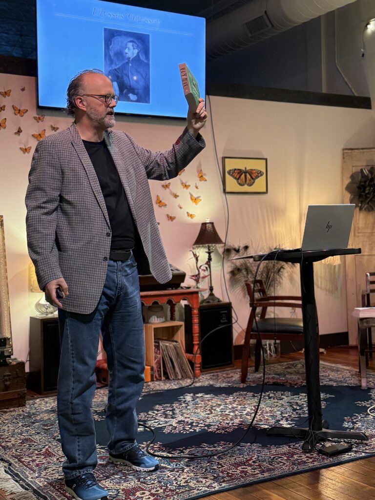 A man in a plaid blazer and jeans holds up a book titled "Ulysses" in front of a projector screen displaying "Ulysses Odyssey" and a portrait.