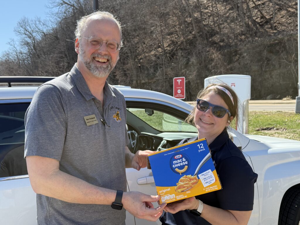 A man and a woman stand next to a white SUV, holding a box of Kraft Mac & Cheese. The man, wearing a gray polo shirt with a name tag that reads "Steve Drahosal, Candidate for County Attorney," smiles at the camera. The woman, wearing sunglasses and a headband, holds the box of mac and cheese. In the background, a Tesla charging station and trees are visible.