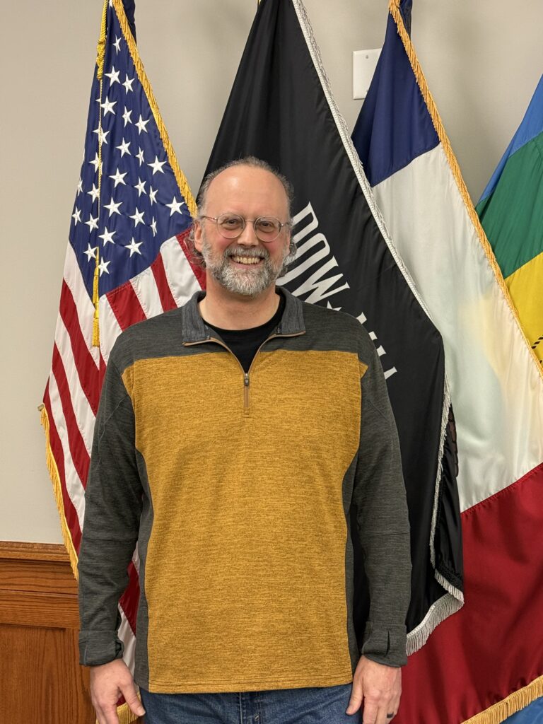 A man with glasses and a beard smiles at the camera, standing in front of several flags, including the American flag and a POW/MIA flag.