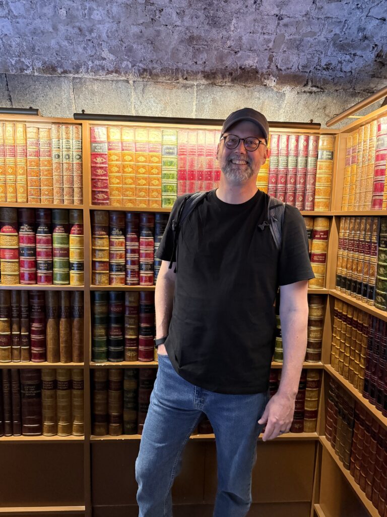 A man wearing a black t-shirt, blue jeans, glasses, and a baseball cap smiles at the camera while standing in front of shelves filled with antique books.