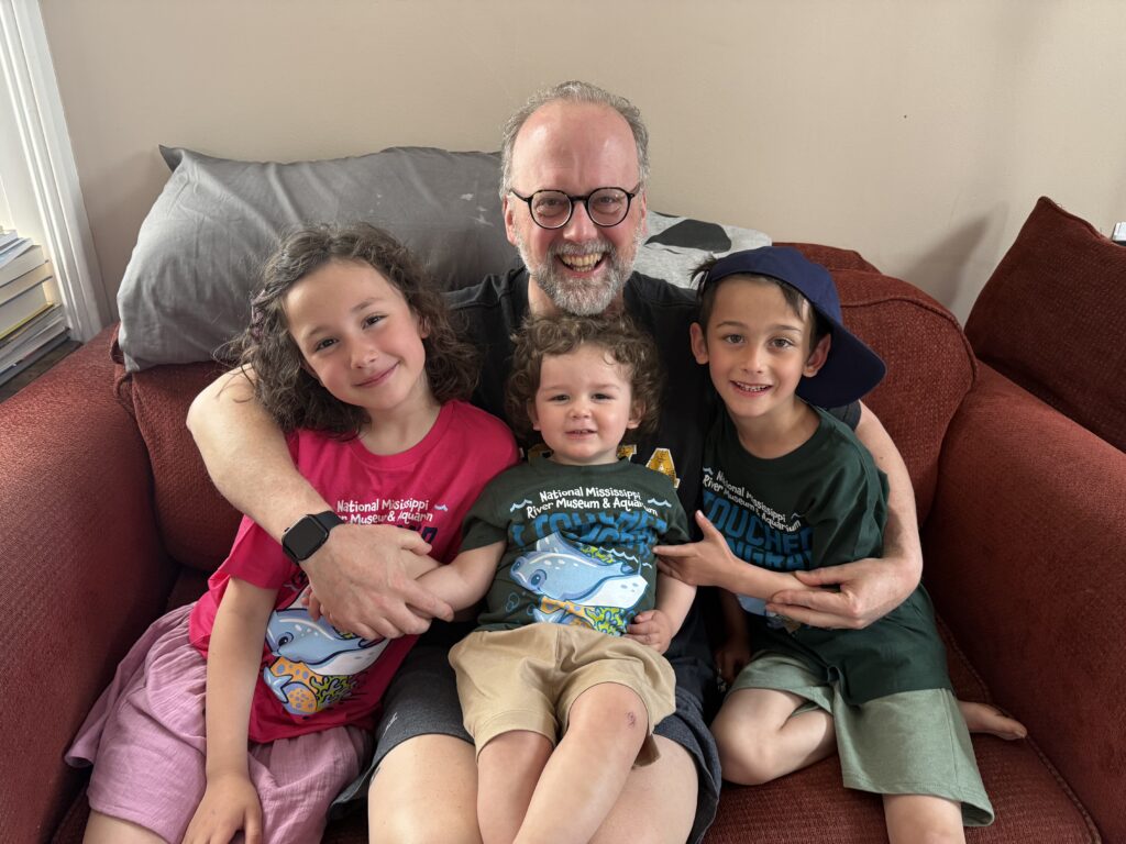 A man smiles while sitting on a red couch with three children. The man is wearing glasses and a black t-shirt. A girl in a pink t-shirt with "National Mississippi River Museum & Aquarium" written on it is on his left. A toddler in a green t-shirt is in the middle, and a boy in a green t-shirt and a blue baseball cap is on his right.