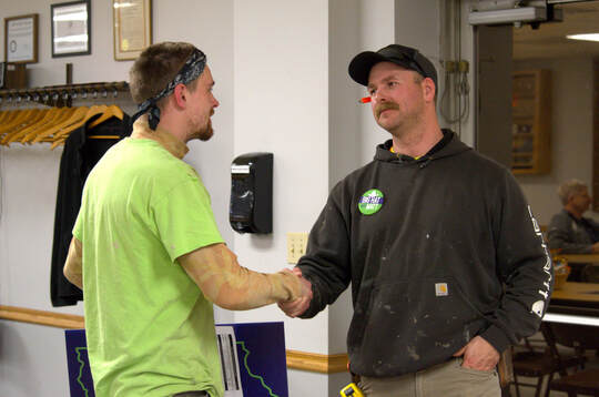 Two men in work clothes shake hands in front of a wall-mounted dispenser.