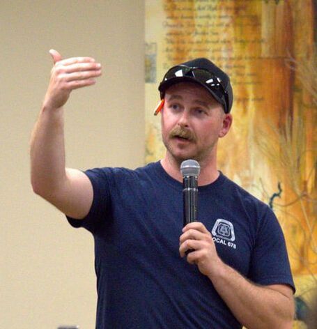 A man wearing a blue t-shirt and a baseball cap with sunglasses on it, holds a microphone and gestures with his left hand.