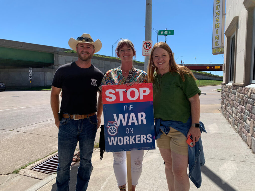 Three people hold a sign that reads "STOP THE WAR ON WORKERS" while standing on a sidewalk.