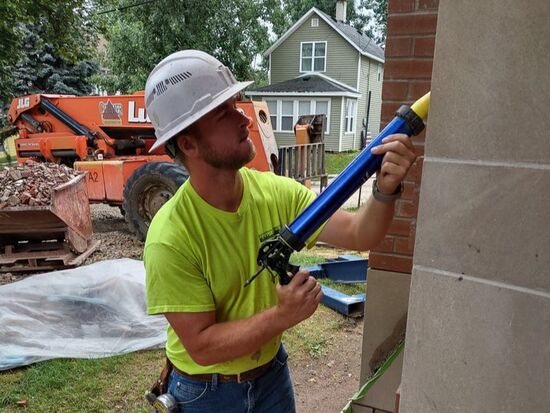A construction worker in a hard hat and neon green shirt uses a caulking gun to seal a stone wall.