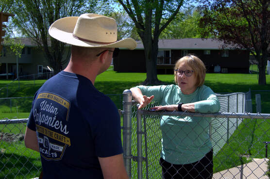 A man wearing a cowboy hat and a navy blue "Union Carpenters" t-shirt talks to a woman wearing glasses and a teal shirt, who is leaning on a chain-link fence.