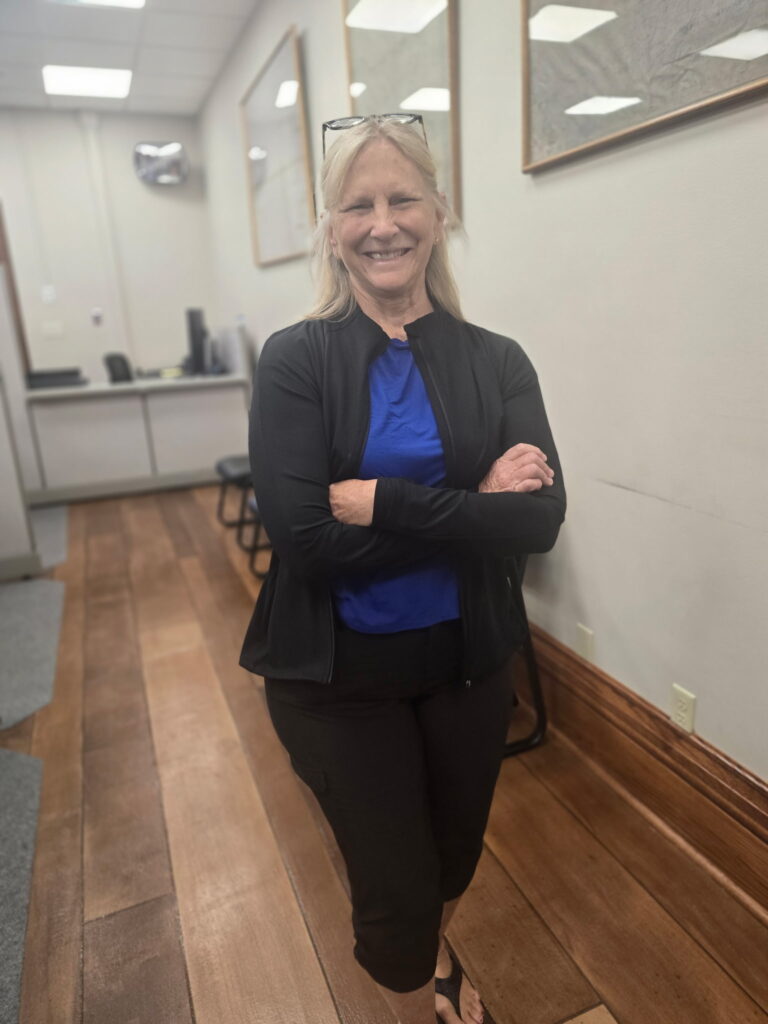 A blonde woman wearing a blue shirt and black jacket smiles with her arms crossed in an office hallway.
