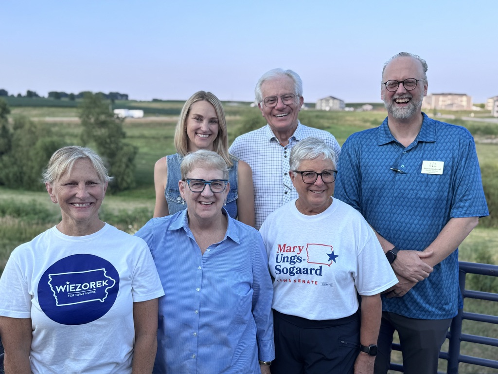 A group of six people, four women and two men, pose for a photo outdoors. The two women in the front are wearing t-shirts with political campaign slogans. The woman on the left wears a white t-shirt with a blue circle and the text "WIEZOREK FOR IOWA HOUSE". The woman in the middle wears a white t-shirt with red and blue text that reads "Mary Ungs-Sogaard IOWA SENATE DISTRICT 42". The other