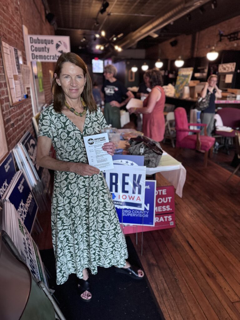 A woman in a green floral dress holds a pamphlet while standing in front of campaign signs for "Rick Dickinson" and "Trek Iowa." A sign for "Dubuque County Democrats" is visible in the background.