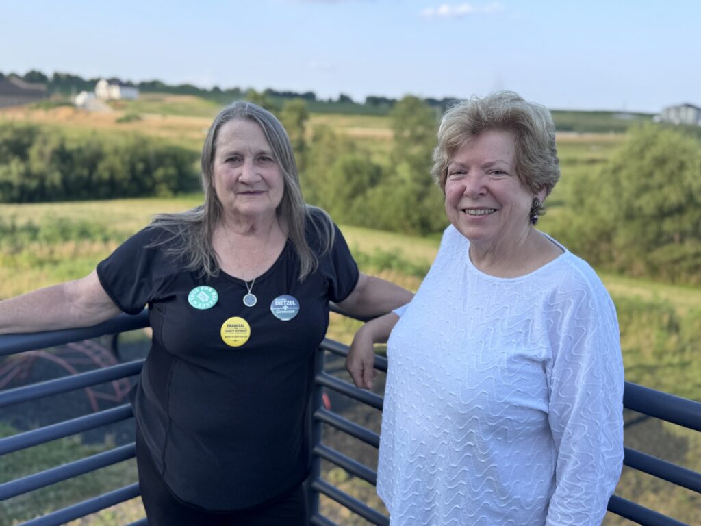 Two women stand on a balcony with a scenic backdrop. The woman on the left wears a black shirt with several campaign buttons, including one that reads "VOTE FOR DEBORAH DRAHOZAL COUNTY ATTORNEY Justice is a full-time job" and another that says "KRISTIN DIETZEL SUPERVISOR". The woman on the right wears a white textured shirt.
