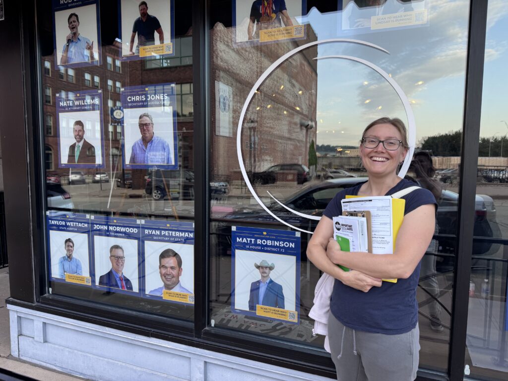 A smiling woman holds campaign materials in front of a storefront window displaying posters of Democratic candidates for Iowa offices.