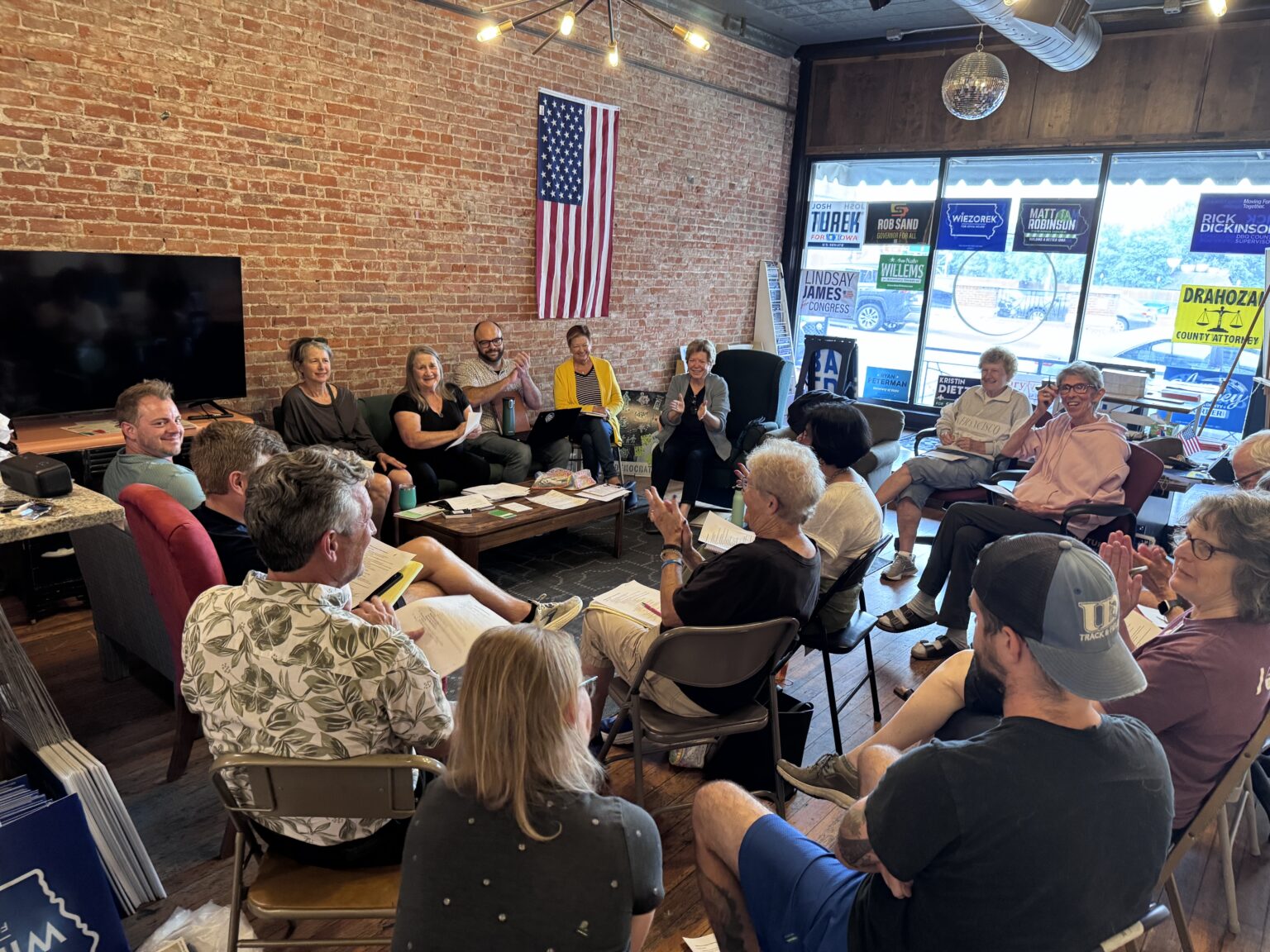 A group of people are gathered in a room with exposed brick walls, likely for a political campaign meeting. There are several campaign signs visible in the background, including "JOSH TUREK FOR IOWA U.S. SENATE", "ROB SAND GOVERNOR FOR ALL", "MATT ROBINSON BUILDING A BETTER IOWA", "RICK DICKINSON COUNTY SUPERVISOR", and "DRAHOZAL COUNTY ATTORNEY". An American flag hangs on the wall. The people are seated in