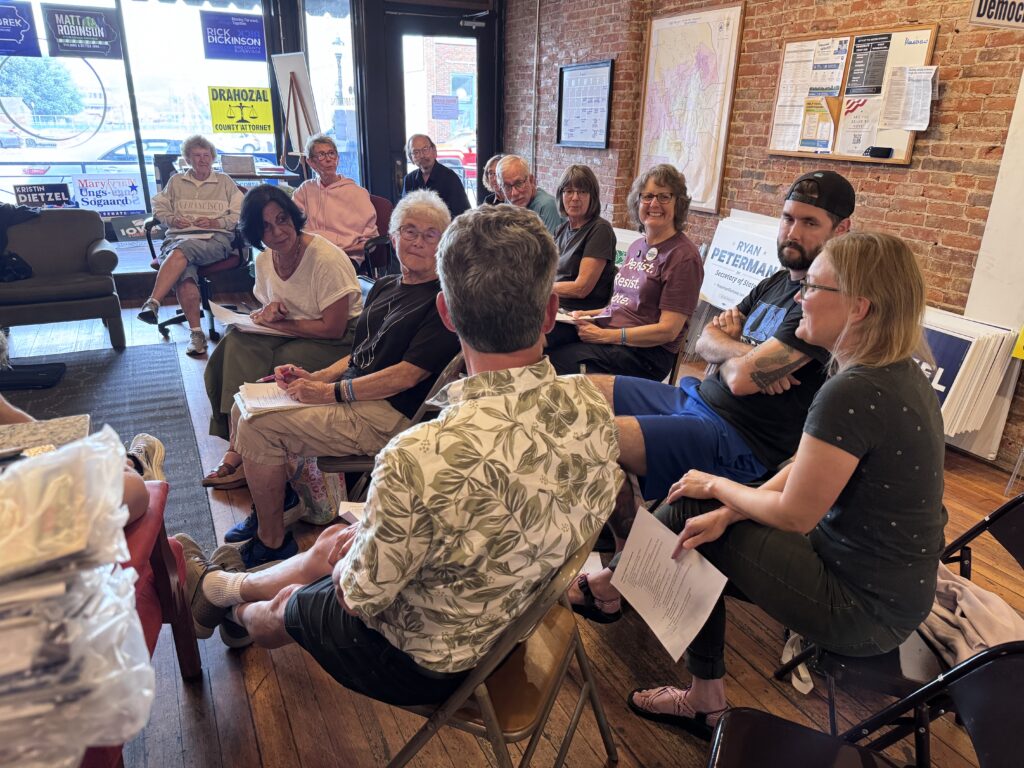 A diverse group of people are seated in chairs in a room with brick walls, attending what appears to be a political meeting or rally. Campaign signs for various candidates, including "MATT ROBINSON FOR IOWA HOUSE," "RICK DICKINSON SUPERVISOR," "MARY UNGS-SOGAARD SENATE," and "RYAN PETERMAN for Secretary of State," are visible in the background and on display. Some individuals are holding papers and listening attentively.