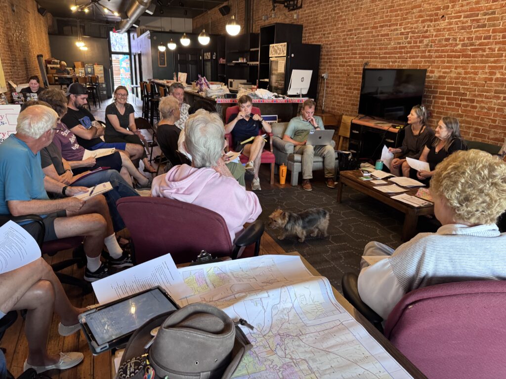 A diverse group of people are gathered in a room, possibly for a meeting or event. Some are seated, while others are standing. A small dog is walking on the floor. There are papers and maps spread out on tables, suggesting a discussion or planning session is taking place. The room has exposed brick walls and industrial-style lighting.