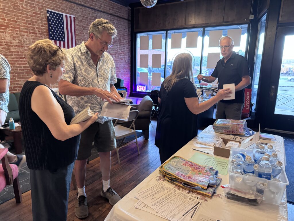 People gather around a table covered in papers and water bottles, with an American flag hanging on a brick wall behind them.