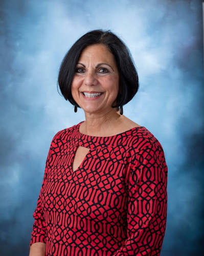 A woman with dark, shoulder-length hair and a red and black patterned shirt smiles at the camera.