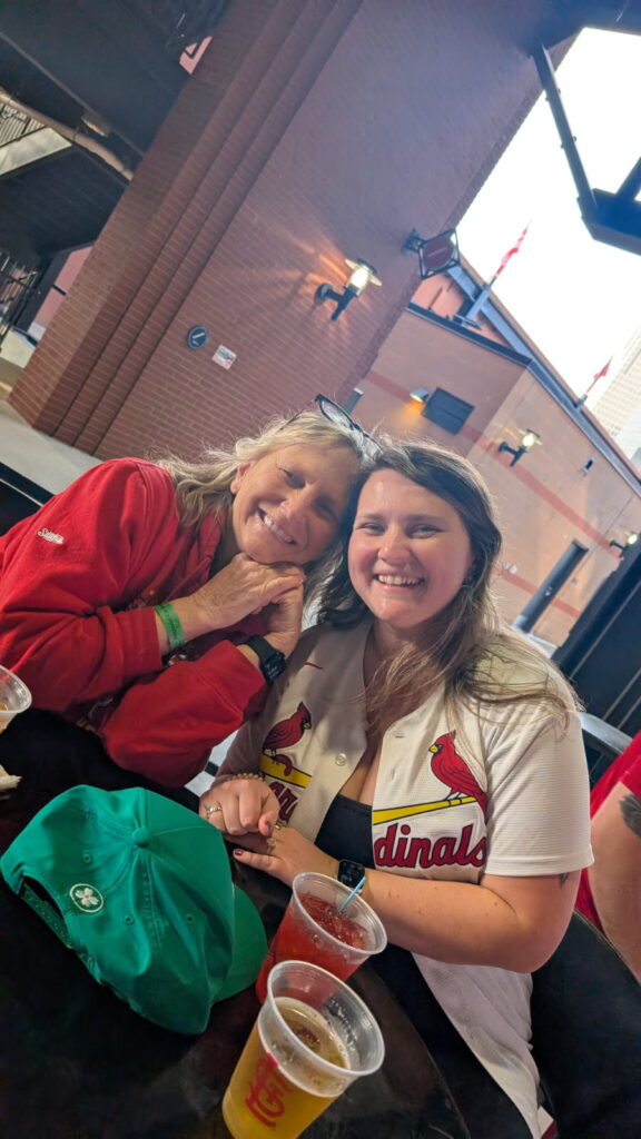 Two smiling women, one wearing a red Cardinals jacket and the other a white Cardinals jersey, pose for a photo at a table with drinks and a green hat.